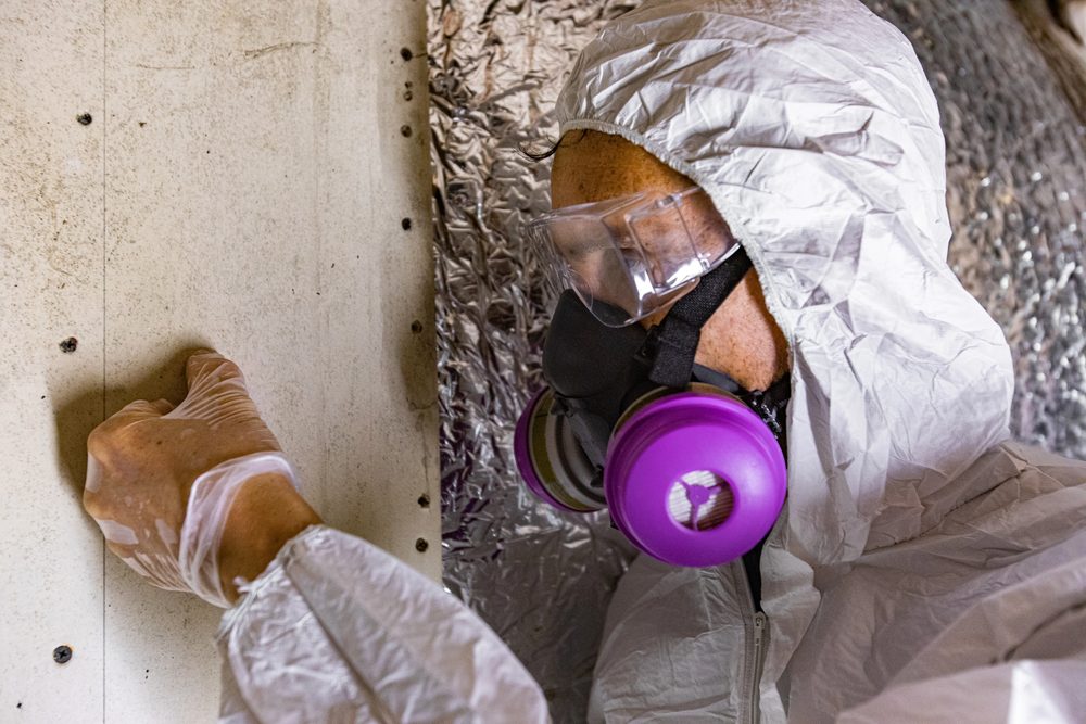 Mold remediation specialist in white hazmat suit, respirator, and goggles inspecting a wall inside a duct space