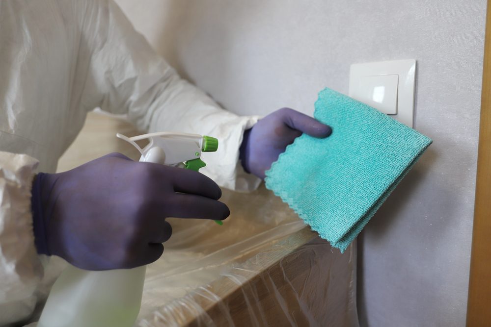 Mold inspector in protective suit and gloves wiping light switch with teal cloth and spray bottle